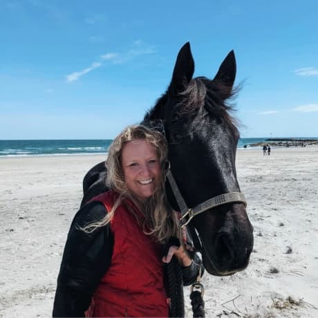 Sara, Medical Team Lead, standing on the beach beside a black horse.